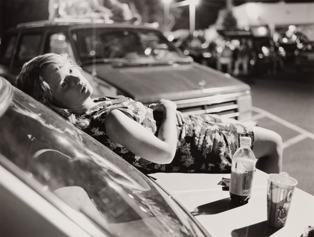 An adolescent girl with short blonde hair reclines on the hood of a car, a bottle of soda and a paper cup filled with ice sit next to her.