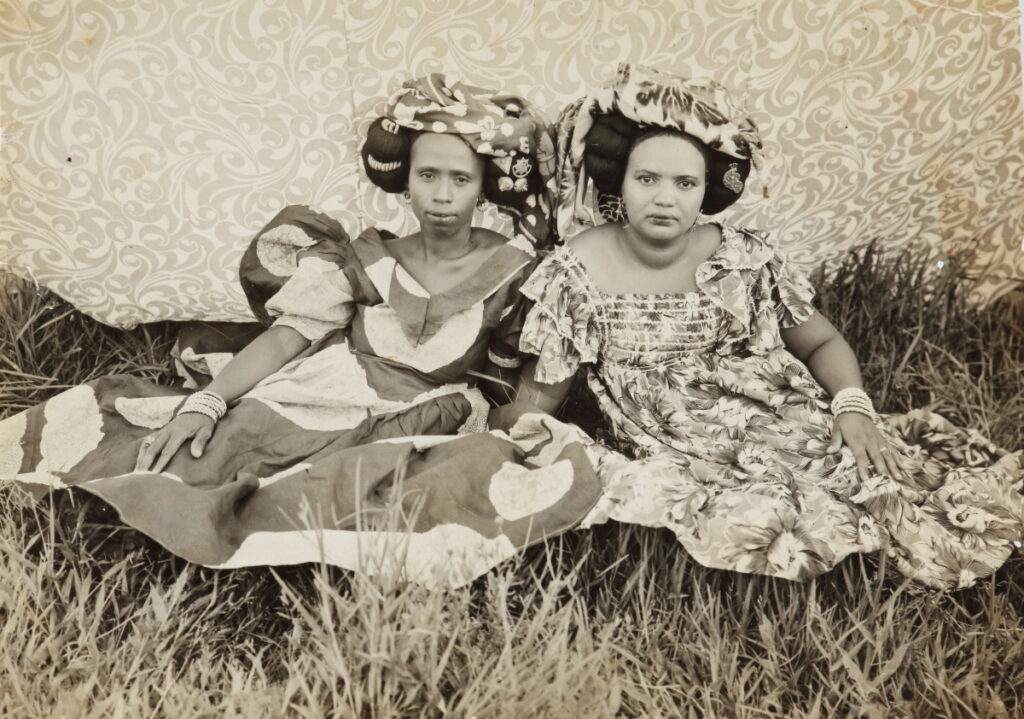 Two women with large hats and dresses in a floral patterns sit in a grassy field with an organic pattern behind them.