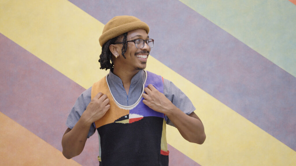 man with dark skin, glasses and a hat smiles while standing in front of a painting with diagonal stripes.
