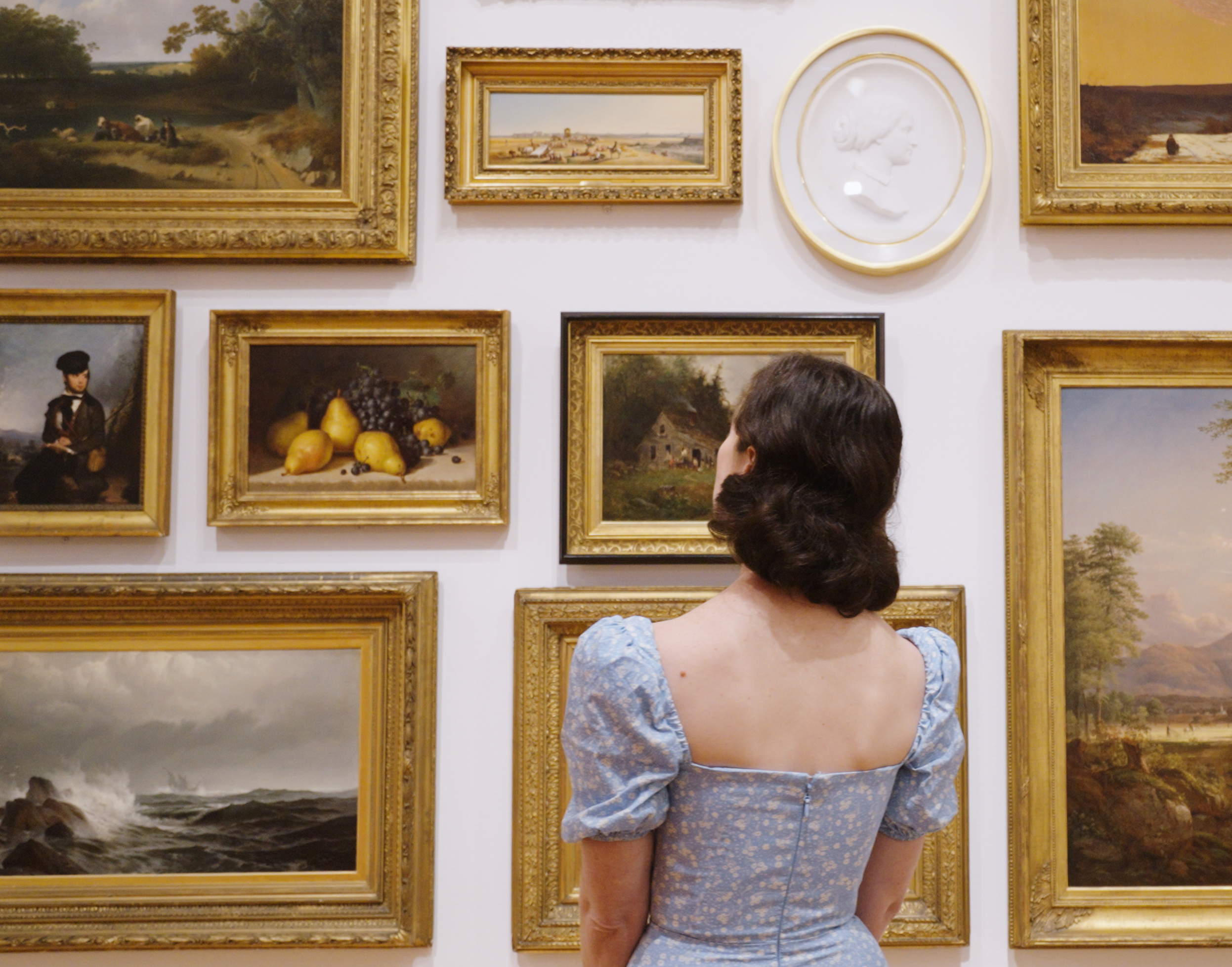 Woman in a blue dress with dark hair looks at a wall with American artworks hung in a salon style.
