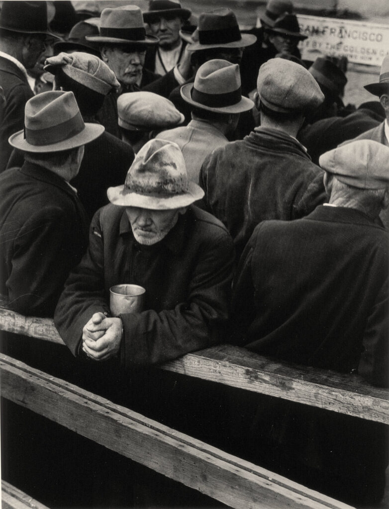 within a crowd, a man in a hat leans against a rail holding a tin can.