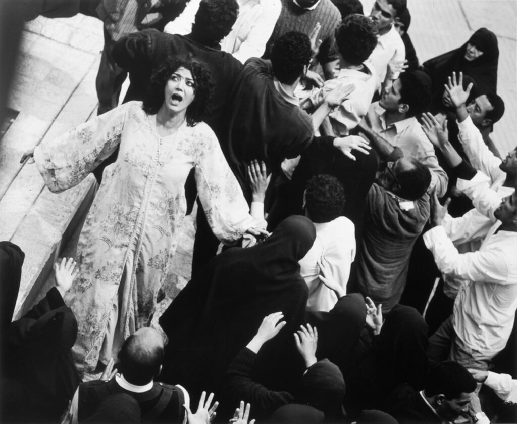 still from a black and white film -- woman in white walks through a crowd of men dressed in black, a disturbed look on her wide-eyed face.