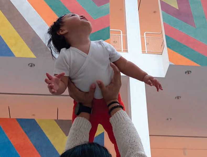 Child is lifted into the air in front of a Sol Lewitt wall painting of rainbow stripes.