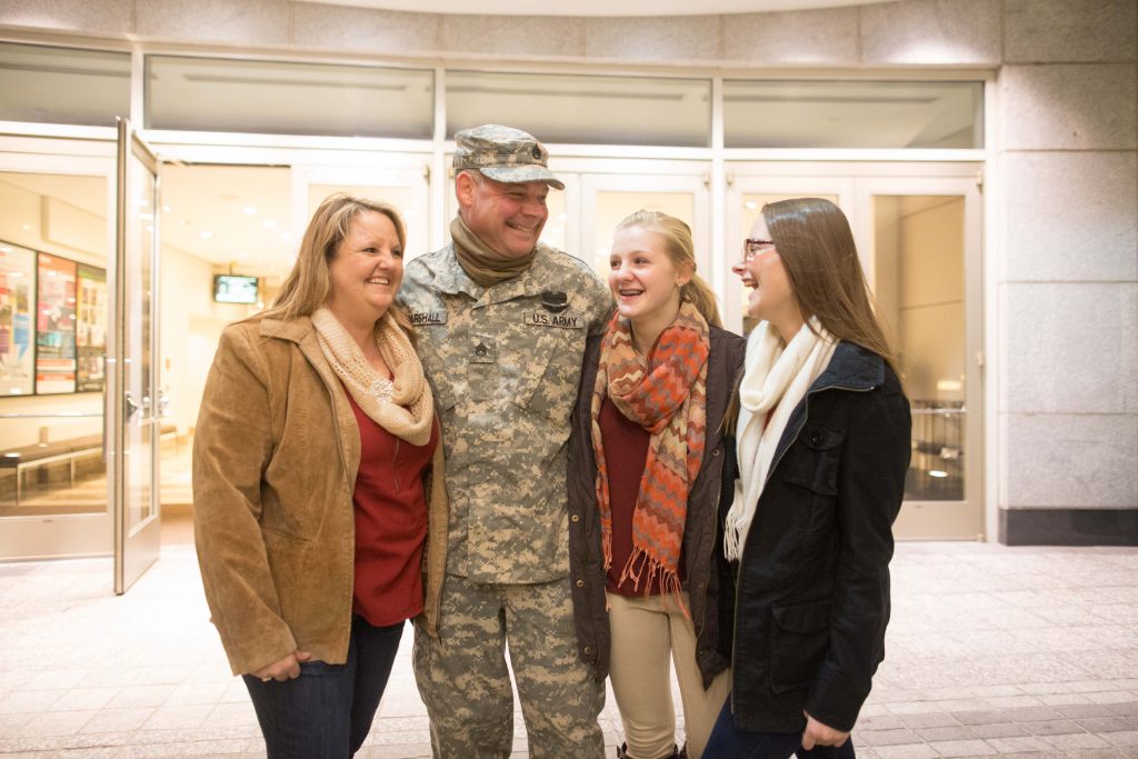 A military service member smiles in his uniform surrounded by his wife and two daughters.