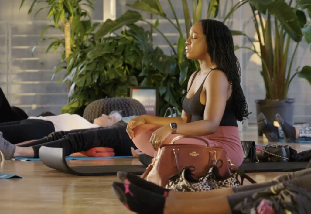 Inside a gallery, a woman with brown skin and long black braids sits cross-legged with her hands on her knees and her eyes closed. Women lay on yoga mats around her.